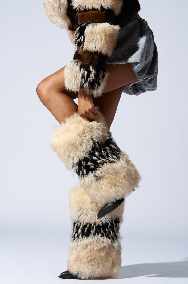 A person poses against a plain background in a stylish outfit with faux fur leg warmers, a cropped furry top, and a mini skirt, all harmonized with beige and brown fur accents, paired with the AZALEA WANG EKKO MULTI FUR STILETTO BOOT.