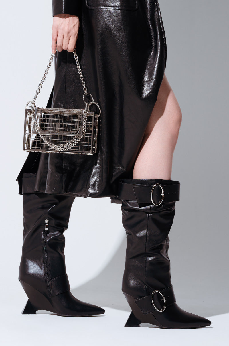 A close-up showcases a person wearing the AZALEA WANG ELABORATE BLACK ABSTRACT WEDGE HEEL BOOT, featuring black leather with large circular buckles and a distinctive wedge heel. The ensemble is completed by a black leather coat, and the person carries a small rectangular handbag with a chain strap against a plain gray backdrop.
