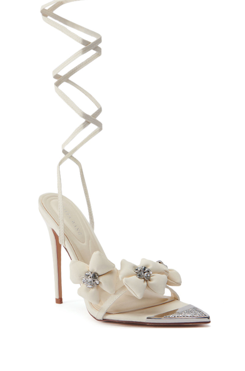 The AZALEA WANG ELAINE EMBELLISHED SANDAL IN TAN is a stylish sandal featuring an open pointed toe design with silver accents. This tan faux leather sandal boasts decorative bows adorned with rhinestones and long laces that wrap around the ankle. The heel is thin and stiletto-style.