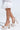 Someone wearing AZALEA WANG ELENAOR Gold Pearl Chunky Sandals with high block heels and ankle straps, paired with a white lace garment featuring a ruffled hem. The faux leather upper subtly complements the outfit against the plain white background.