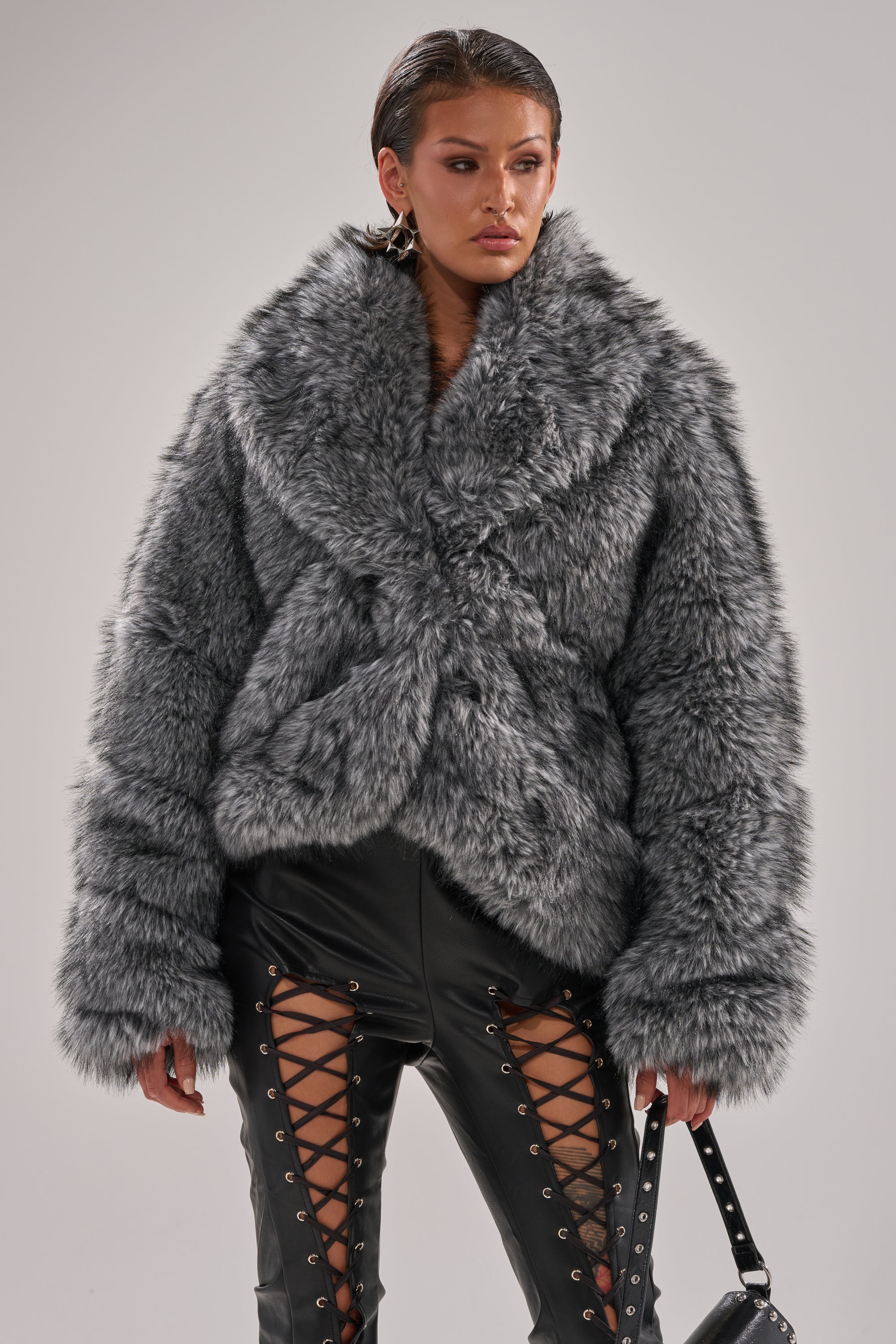 A woman stands against a plain background in the ELIXIR PANELED FAUX FUR IN GREY jacket with an oversized collar, black lace-up leather pants, and holds a black studded purse in her left hand. 2