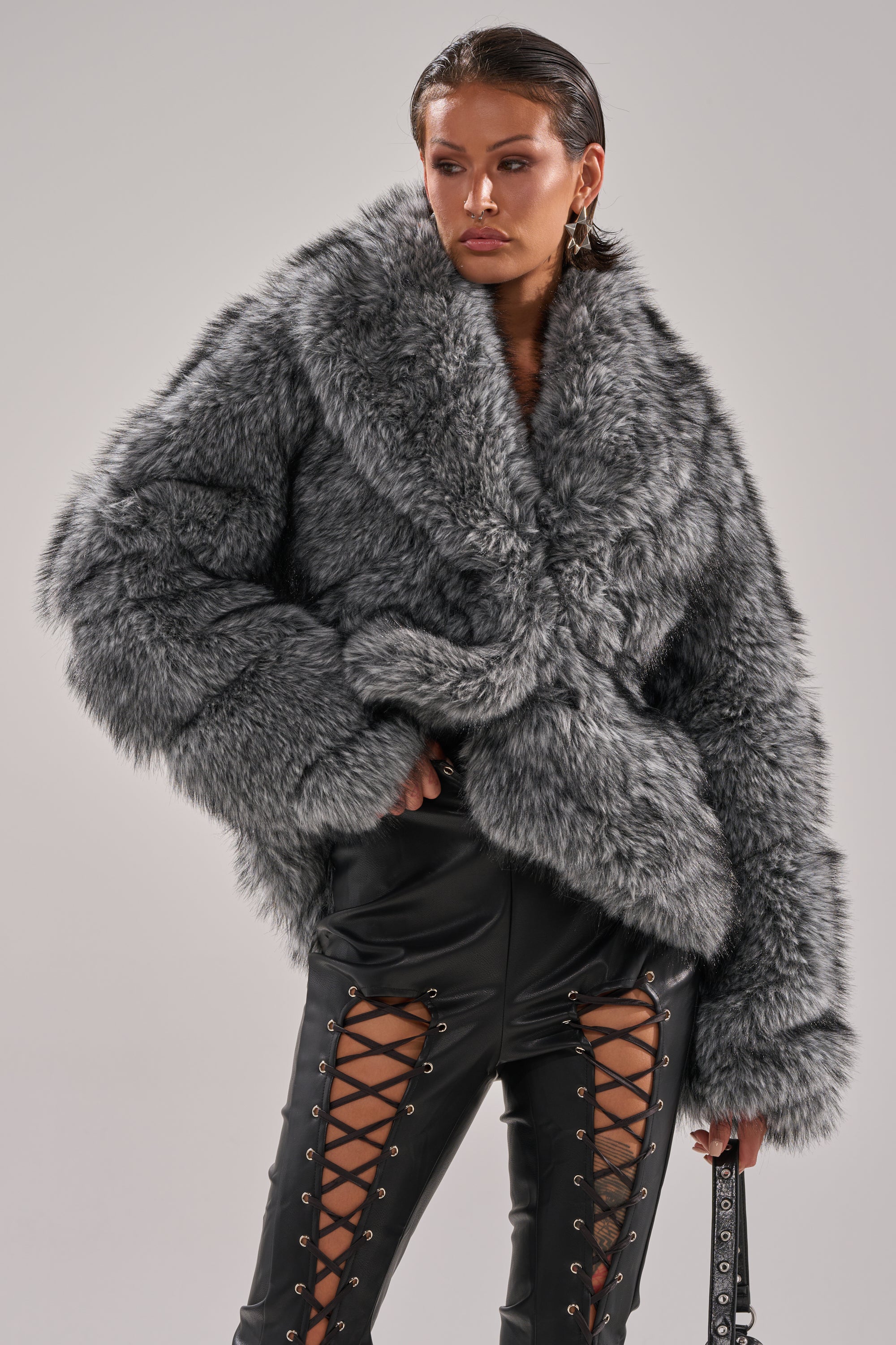 A person stands confidently against a plain background wearing the ELIXIR Paneled Faux Fur in Grey paired with black lace-up front leather pants. 3