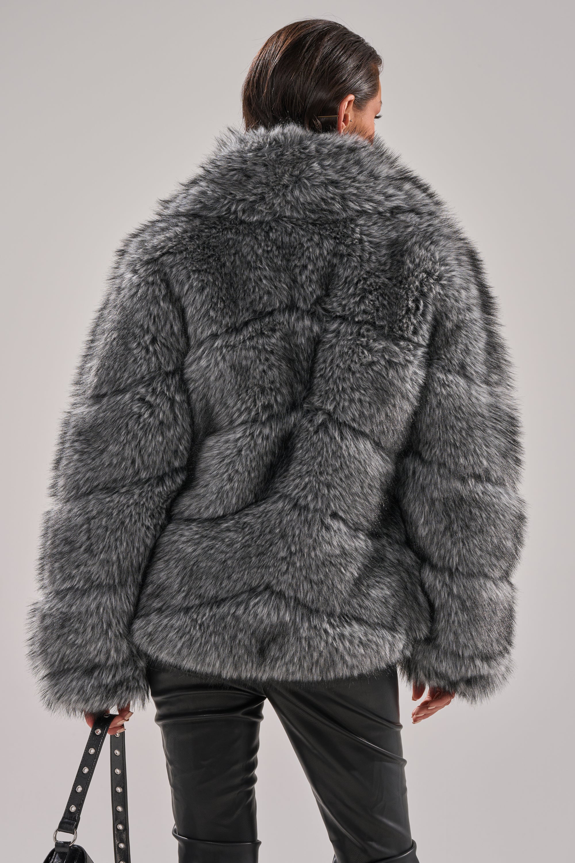 A person with short dark hair, seen from the back, wears the ELIXIR PANELED FAUX FUR IN GREY coat with black leather pants and holds a studded black bag against a plain light background. 5