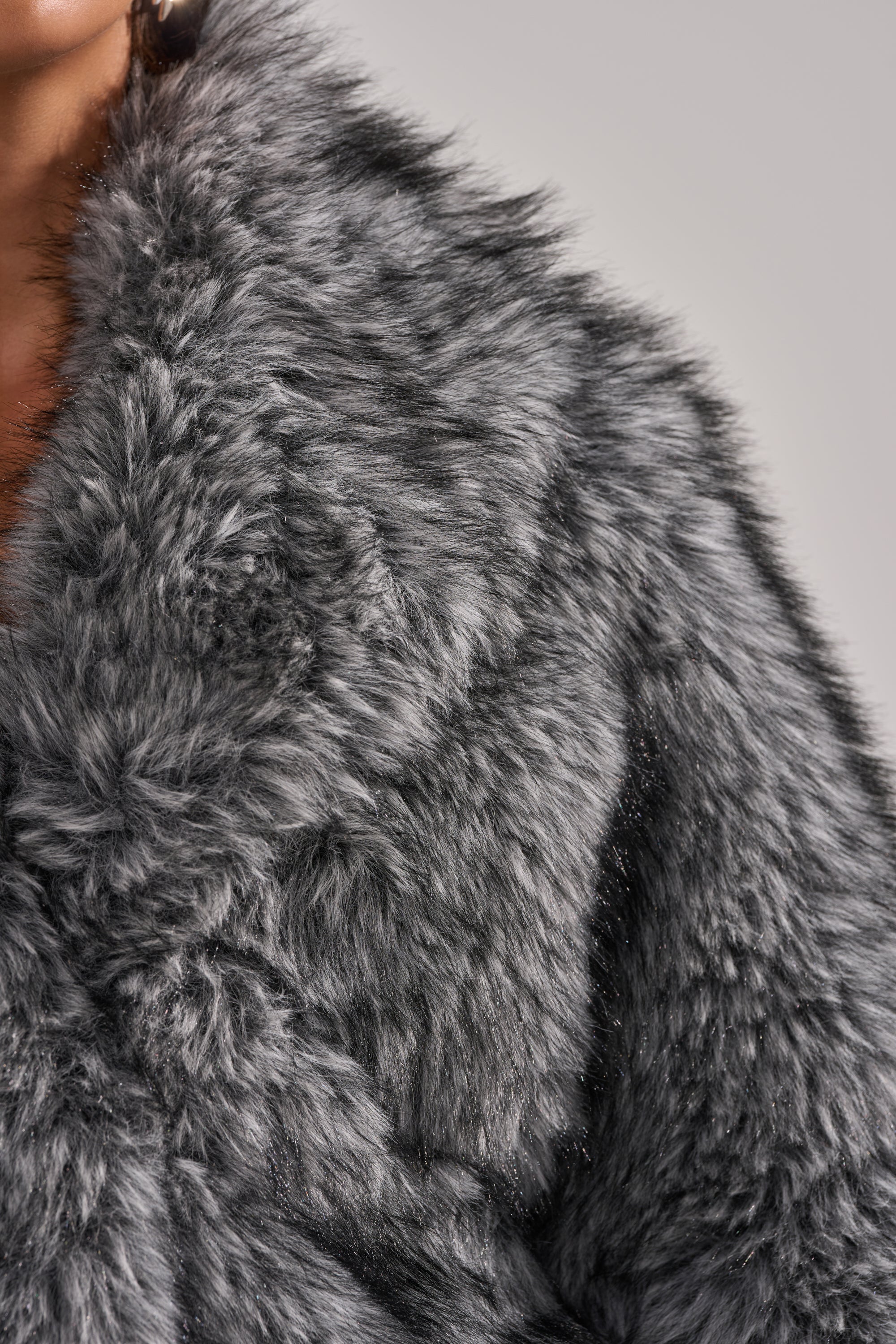 Close-up of a person wearing the ELIXIR PANELED FAUX FUR IN GREY, featuring an oversized collar and plush texture that exudes cozy elegance. The thick gray faux fur stands out against a neutral background, with only part of the face visible. 8