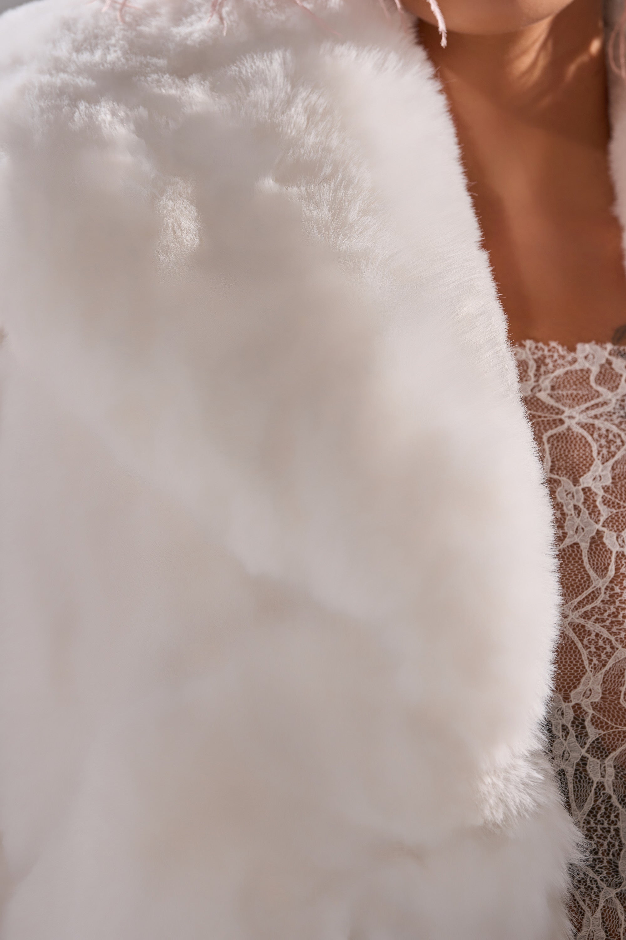 Close-up of a person wearing the ELIXIR PANELED FAUX FUR IN WHITE, styled as a chic winter coat, draped over a beige lace garment with part of their neck and shoulder visible.