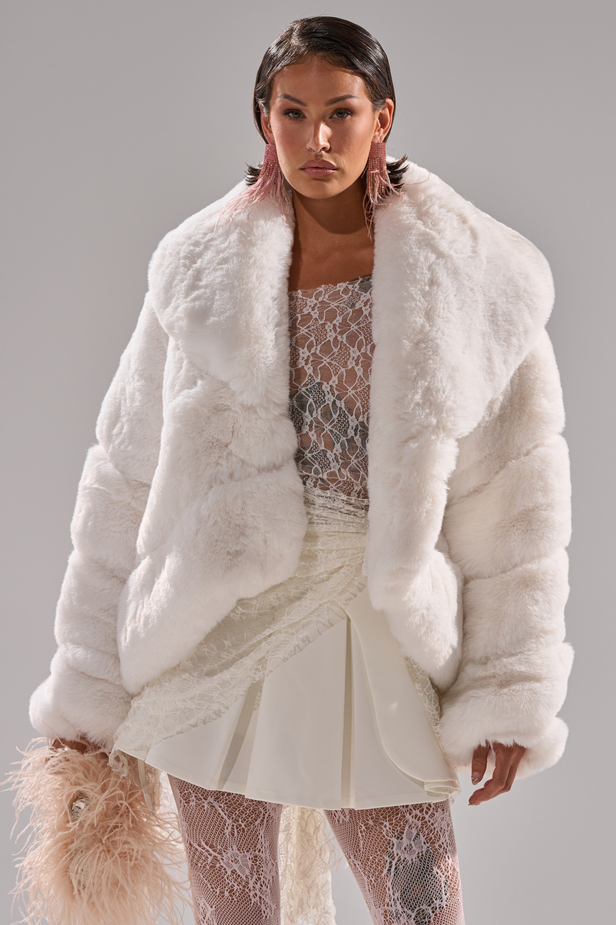 A woman wears the ELIXIR PANELED FAUX FUR IN WHITE over a sheer lace dress with a white mini skirt overlay, holding a feathered accessory. She has slicked-back short hair and stands against a plain gray background.