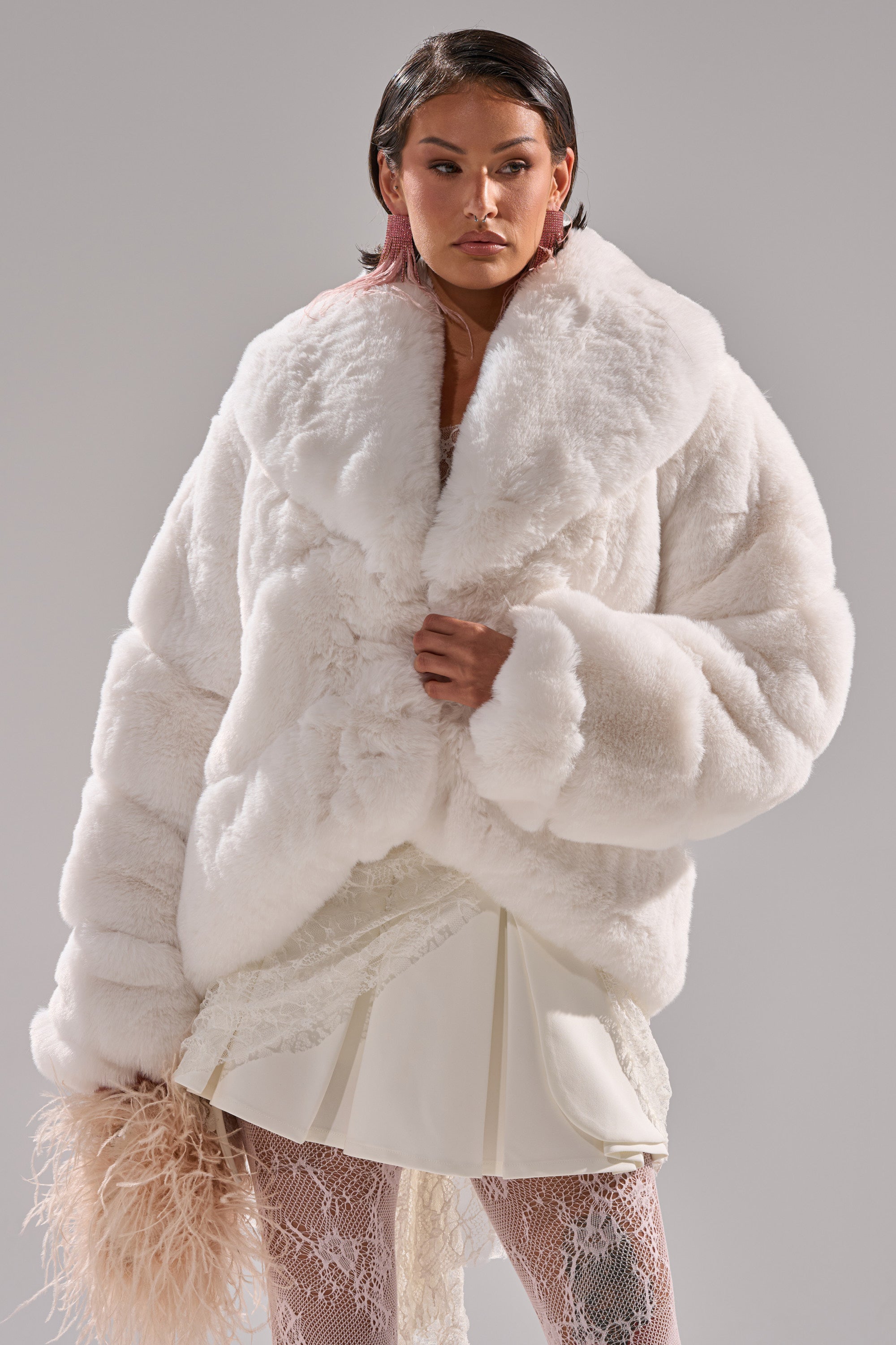 A woman stands confidently against a plain gray background in the ELIXIR Paneled Faux Fur in White, styled with a short white skirt, white lace tights, and holding a light furry accessory.