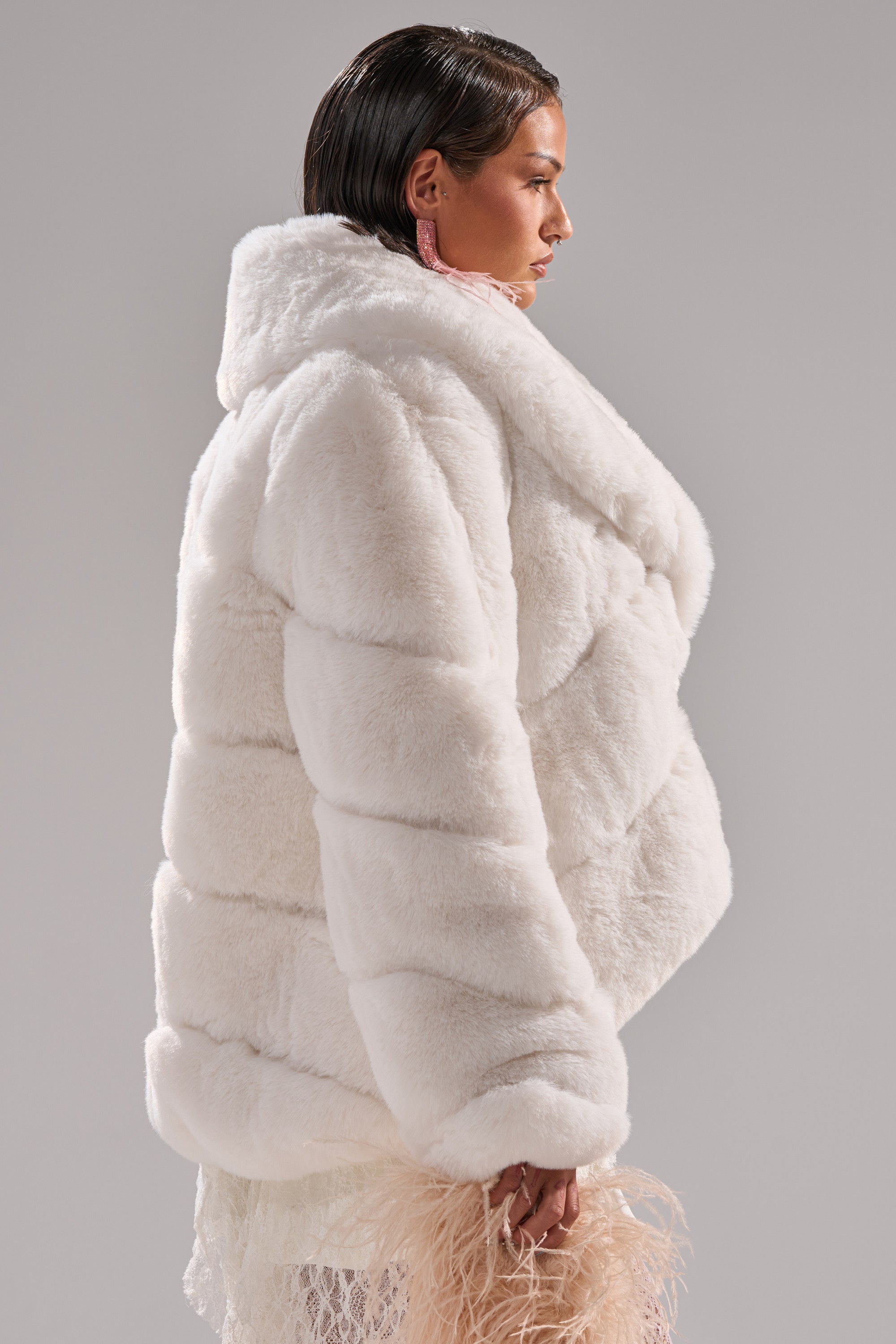 A woman with short, slicked-back hair wears the ELIXIR PANELED FAUX FUR IN WHITE and holds a pale pink feathered accessory, standing in profile against a neutral gray background.