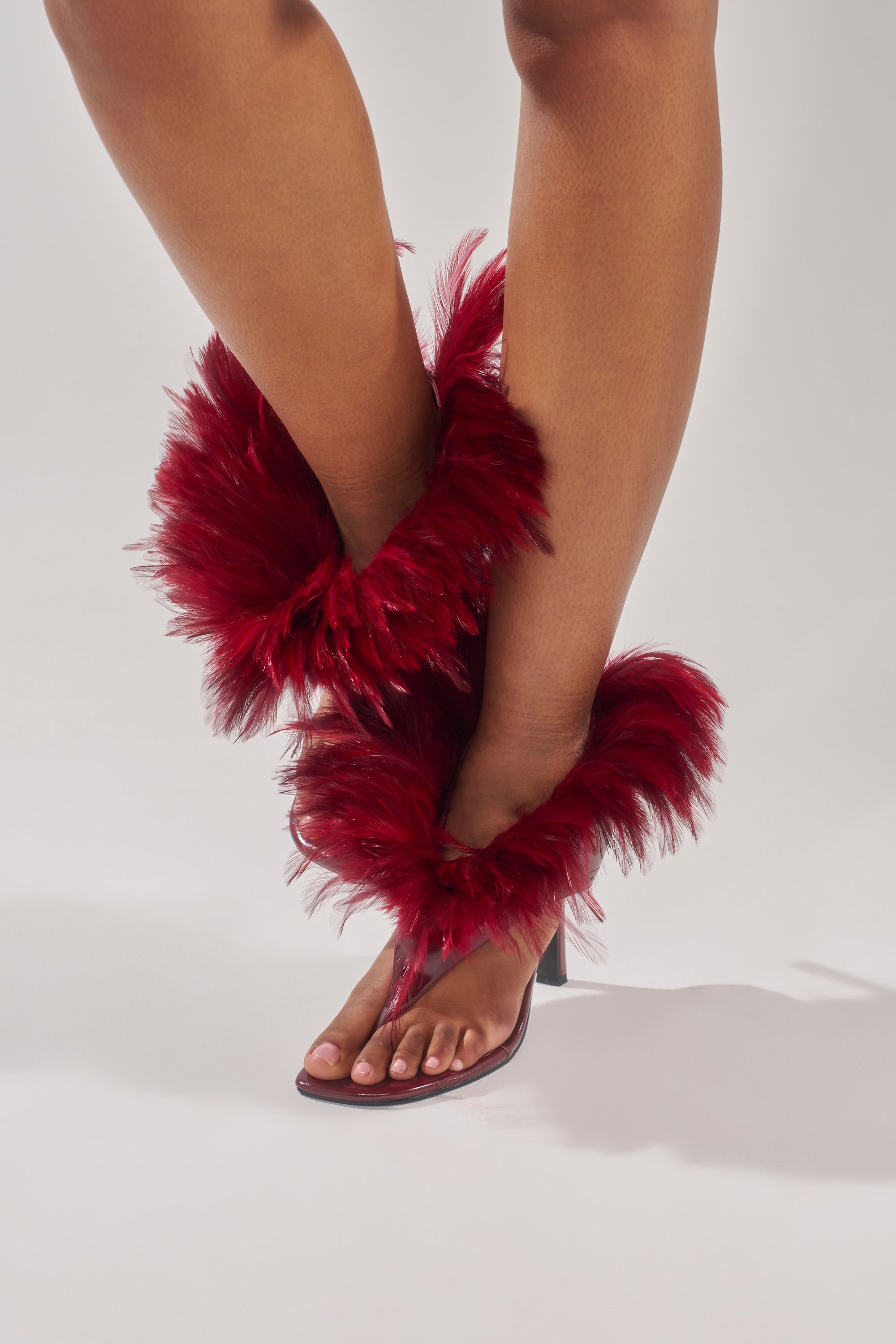 A person stands on a white background wearing the AKIRA GOLD ELIZAVETA BURGUNDY FEATHER THONG SANDAL, featuring stiletto heels and dramatic burgundy feathers that adorn the ankles and lower legs. 1