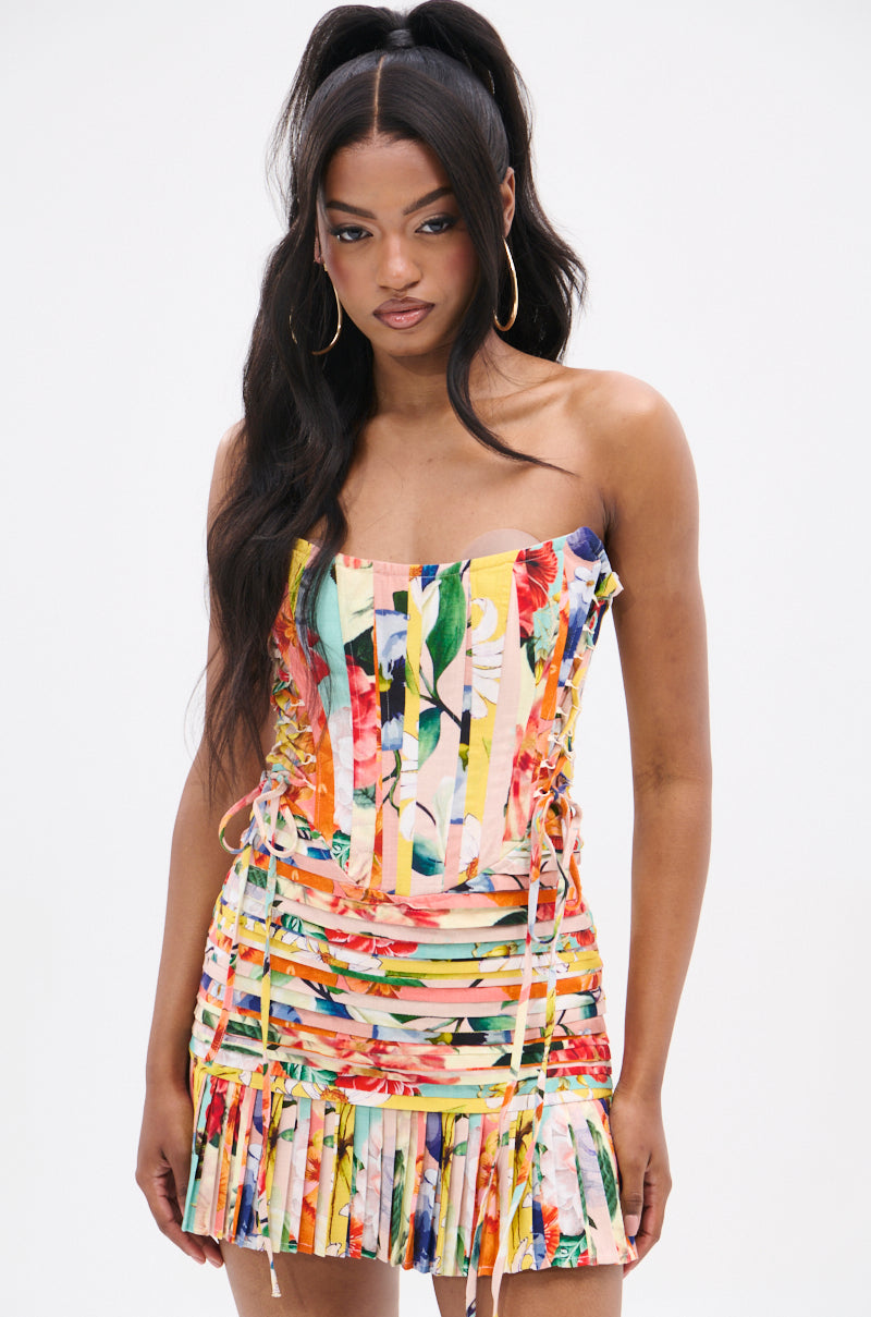 A woman with long dark hair in a high ponytail wears the FLOUNCE CORSET MINI DRESS, featuring a strapless neckline and mixed print. Gold hoop earrings complement her look as she stands against a plain white background.