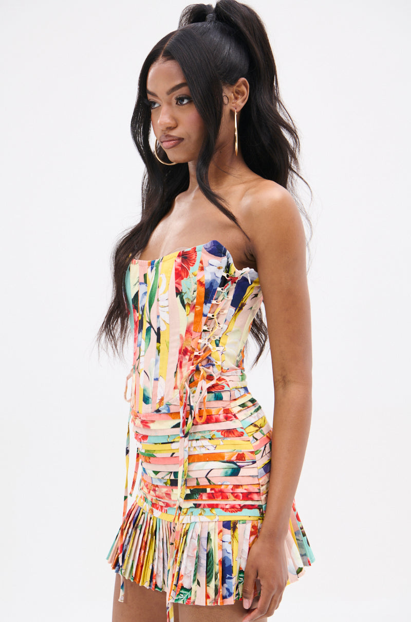 Wearing hoop earrings, a woman with long dark hair stands against a plain white background in the strapless FLOUNCE CORSET MINI DRESS. It features mixed print fabric, a lace-up front, and pleated skirt. She looks to the side.