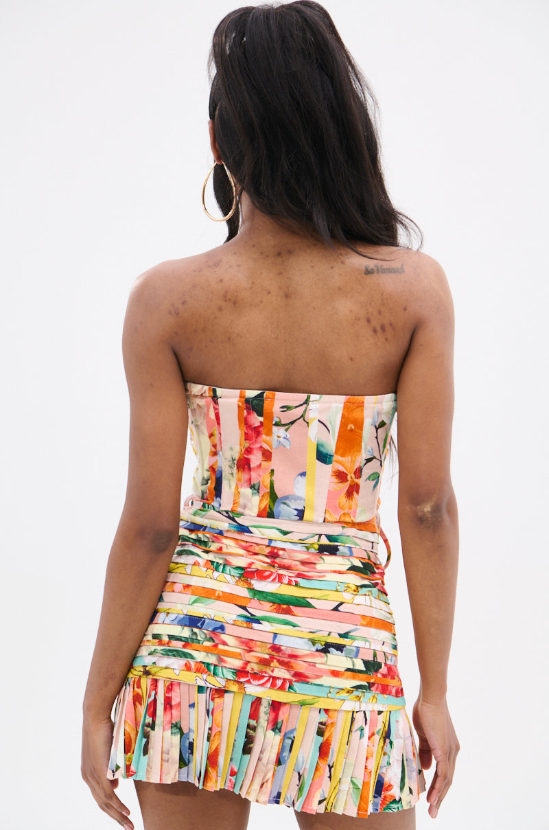A woman with long dark hair is shown from behind, wearing the FLOUNCE CORSET MINI DRESS with a strapless neckline and pleated skirt. She has a small shoulder tattoo, large hoop earrings, and the mixed floral-striped print pops against the plain white background.