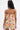 A woman with long dark hair is shown from behind, wearing the FLOUNCE CORSET MINI DRESS with a strapless neckline and pleated skirt. She has a small shoulder tattoo, large hoop earrings, and the mixed floral-striped print pops against the plain white background.