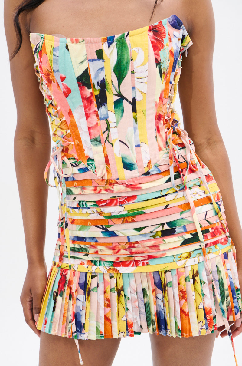 A person wearing the Flounce Corset Mini Dress, featuring a colorful floral strapless neckline, lace-up sides, and a pleated skirt, stands against a plain white background.