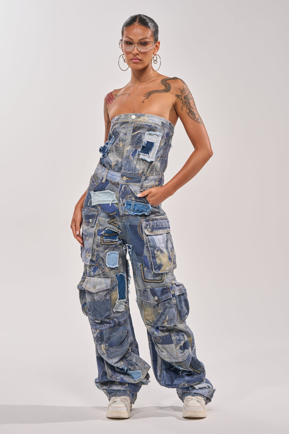 Wearing the PATCHWORK DENIM ON DENIM JUMPSUIT with wide legs and large pockets in mixed denim shades, paired with white sneakers, hoop earrings, clear glasses, and showing arm tattoos against a plain background. 0