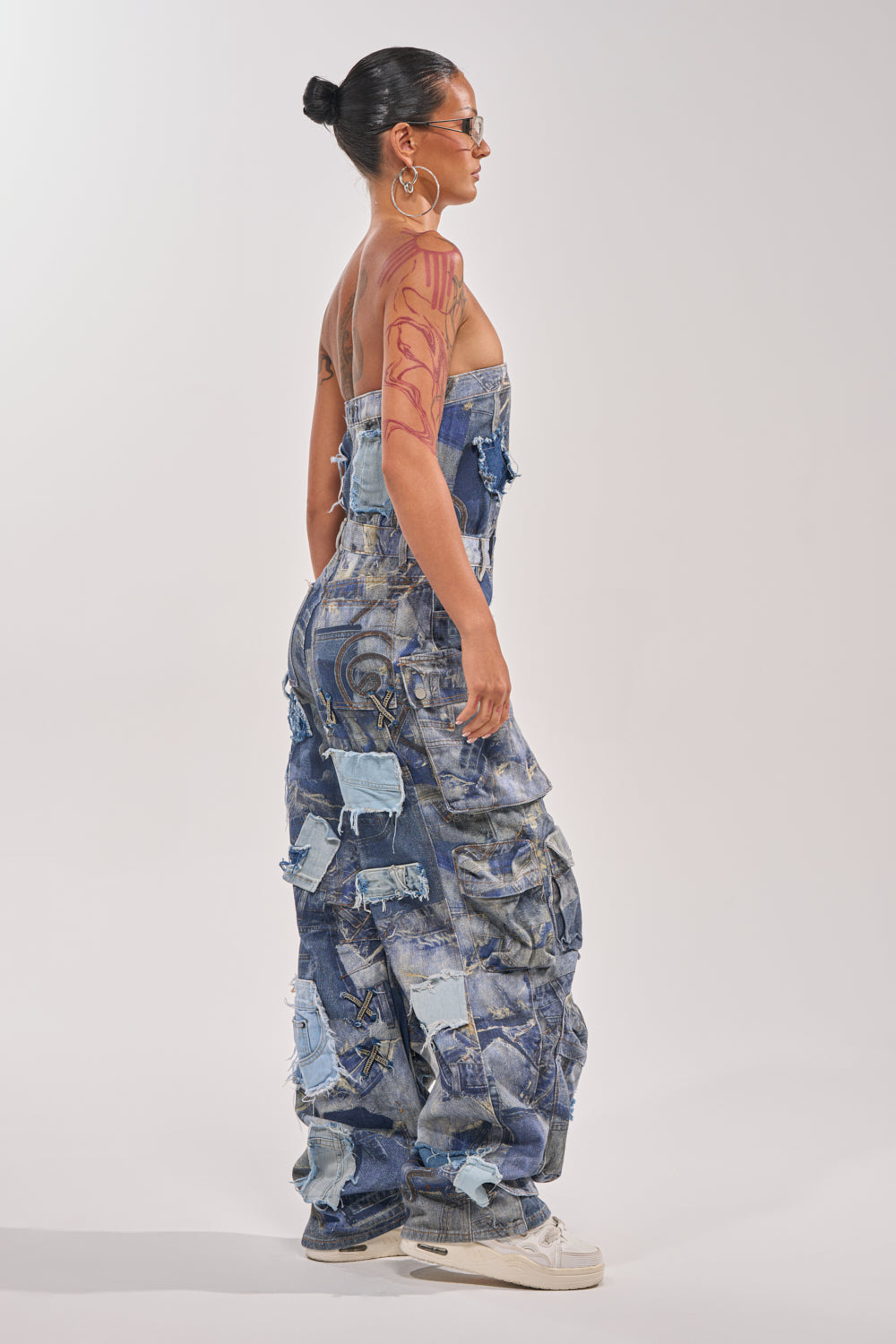 A woman stands sideways wearing the PATCHWORK DENIM ON DENIM JUMPSUIT featuring a strapless, wide-leg silhouette with faded blue patchwork. She pairs it with a bun hairstyle, hoop earrings, sunglasses, red arm tattoos, and white sneakers. 2