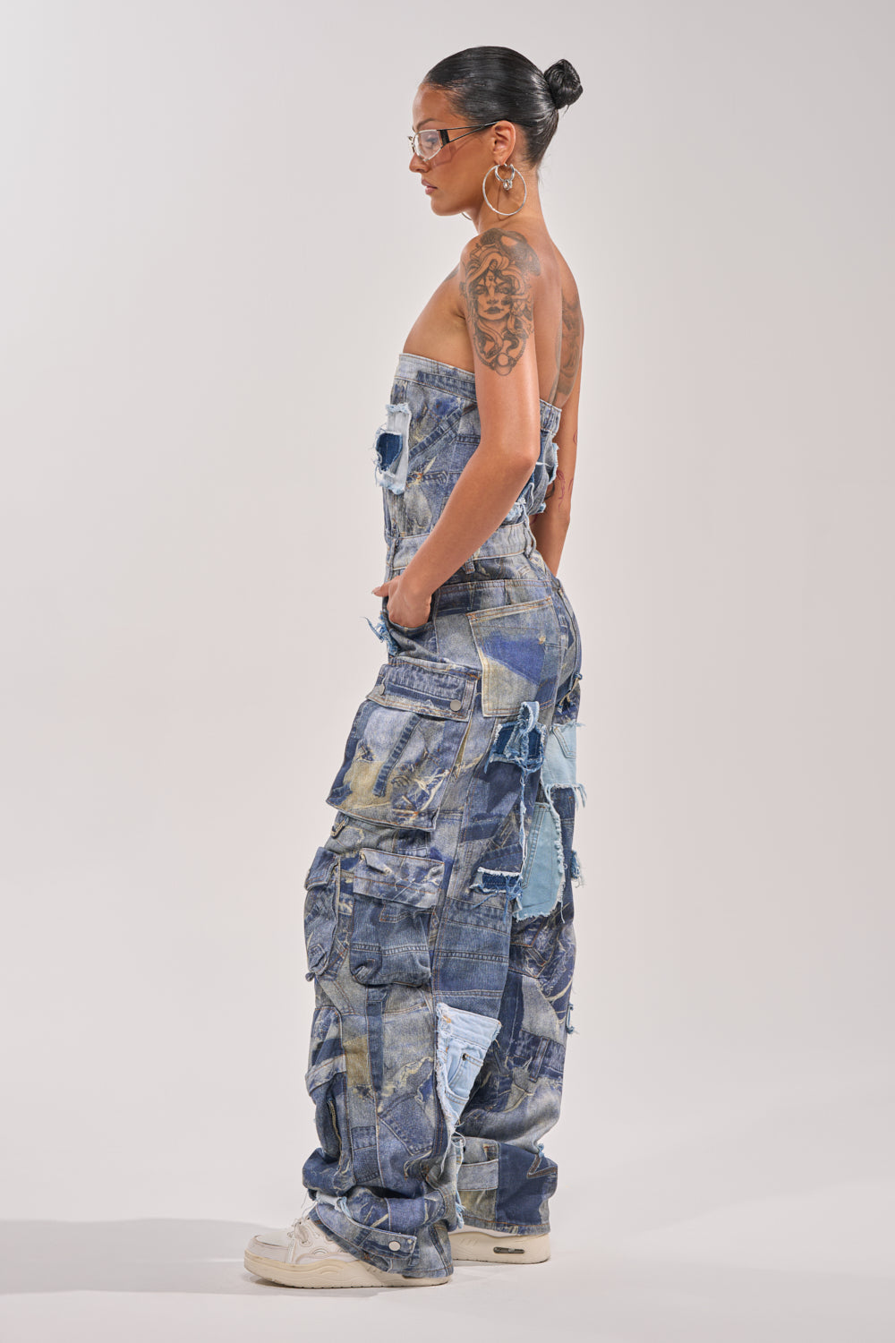 Woman in profile against a plain background, wearing the PATCHWORK DENIM ON DENIM JUMPSUIT with large hoop earrings, glasses, visible arm tattoos, and her hair styled in a neat bun. 3