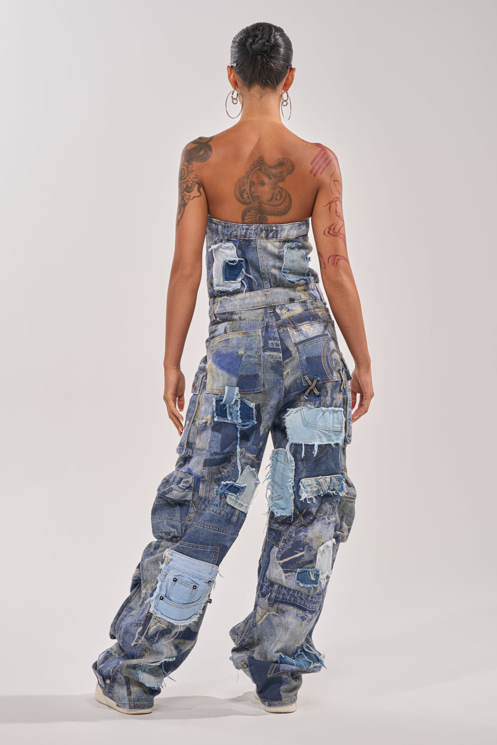 A tattooed individual with large hoop earrings models the PATCHWORK DENIM ON DENIM JUMPSUIT, standing with their back to the camera against a plain, light background. 4
