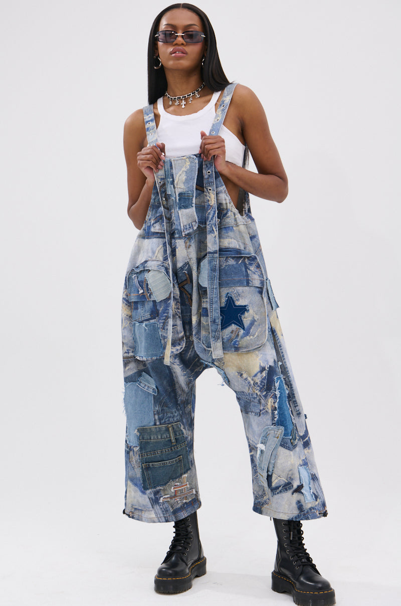 A person stands confidently against a plain background, wearing FOR THE GRAM PATCHWORK OVERALLS with star-patterned patchwork, a white tank top, sunglasses, and black combat boots, holding the adjustable shoulder straps.