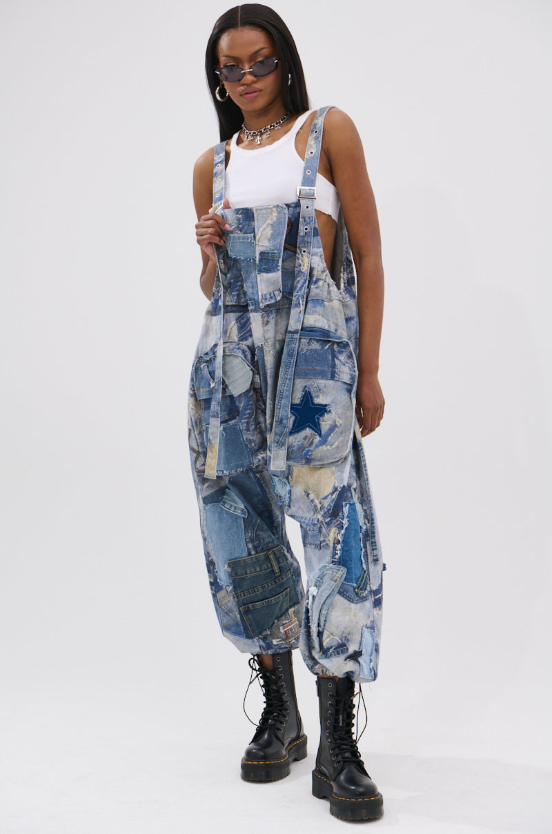 Wearing FOR THE GRAM PATCHWORK OVERALLS with adjustable straps, a blue star detail, a white tank top, black combat boots, sunglasses, and layered necklaces, a person poses against a plain white background.