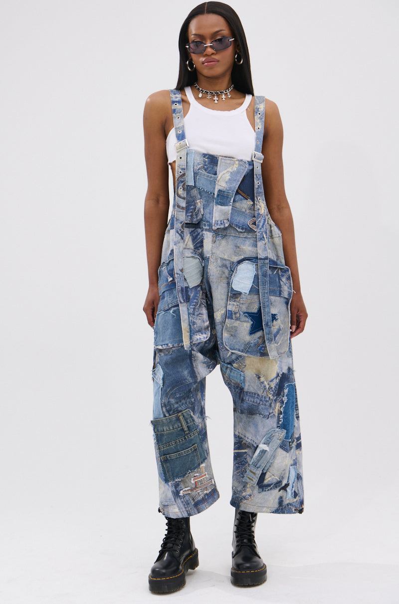 A person models the FOR THE GRAM PATCHWORK OVERALLS—oversized, denim, with adjustable straps—styled with sunglasses, a white tank top, black combat boots, and a silver chain necklace against a plain white background.