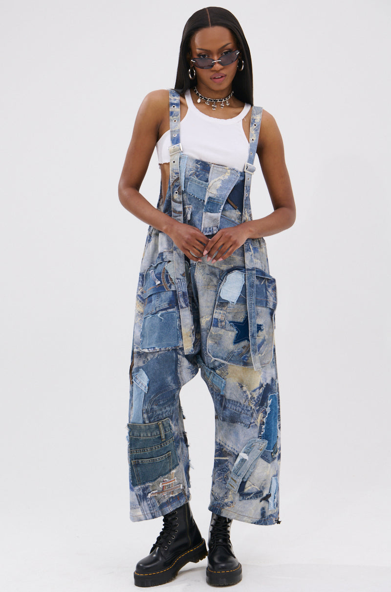 A person wearing FOR THE GRAM PATCHWORK OVERALLS with adjustable straps, a white tank top, sunglasses, necklace, earrings, and chunky black boots stands confidently. Their straight hair complements the bold patchwork denim style.