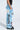 Wearing FIRST CLASS TICKET GRAPHIC CARGO RELAXED LEG JEANS featuring colorful prints and text, a person pairs them with a black top and chunky black shoes, standing with one hand in their pocket against a plain background.