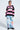 A person wearing glasses, a pink and black striped "Leader" sweater, and FIRST CLASS TICKET GRAPHIC CARGO RELAXED LEG JEANS with colorful patches stands against a plain light background, looking slightly to the side.