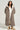 A person wearing the BEAR HUG REVERSIBLE FLEECE TRENCH, featuring a long hood and an oversized silhouette in brown, layered over a light gray outfit. They pair it with tan boots and have long, curly hair while standing against a light, neutral background.