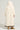 A person wearing the BEAR HUG REVERSIBLE FLEECE TRENCH, a long, hooded coat with an oversized silhouette in cream sherpa material, stands with their back to the camera. The coat features a contrasting dark hem and is paired with tan boots. The background is a plain light color.