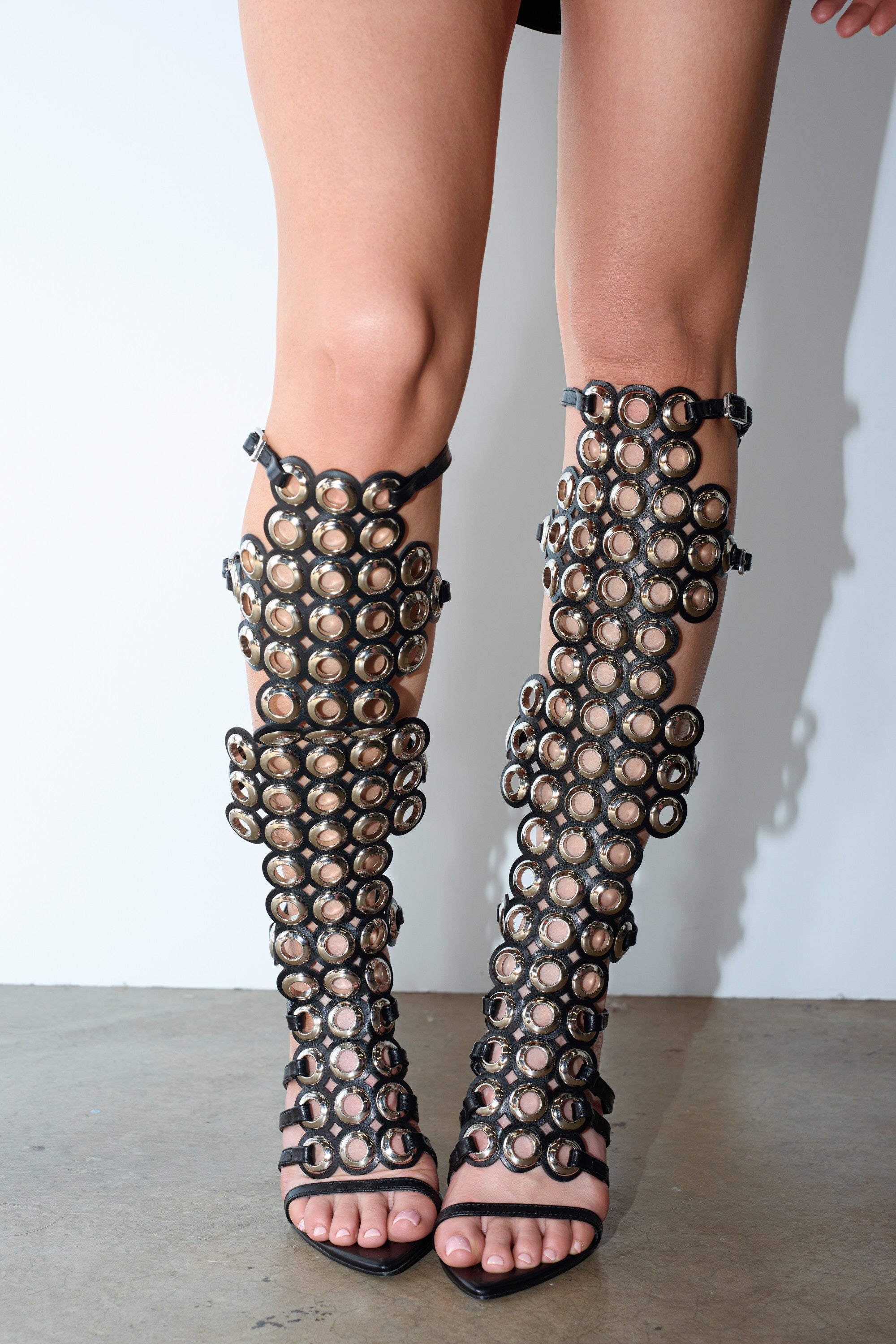 A person wears the AZALEA WANG ELLINOR Black Grommet Detail Gladiator Sandal with knee-high straps, circular cutouts, and metallic accents while standing on a concrete floor against a white wall.