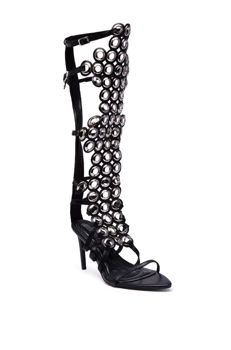 The AZALEA WANG ELLINOR BLACK GROMMET DETAIL GLADIATOR SANDAL is a bold knee-high stiletto with eye-catching grommet studs, open toes, and slender ankle and toe straps for a standout look.