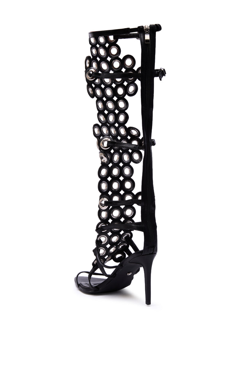 The AZALEA WANG ELLINOR Black Grommet Detail Gladiator Sandal is a knee-high stiletto with circular cutouts, multiple straps, and bold grommets, extending to the knee and displayed on a white background.