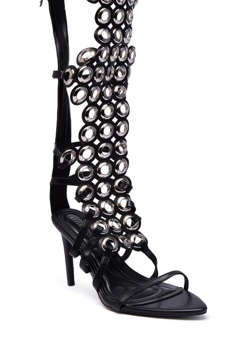 The AZALEA WANG ELLINOR Black Grommet Detail Gladiator Sandal features a high stiletto heel, open toe, strappy lower section, and a tall upper adorned with multiple metallic rings and bold grommet accents.