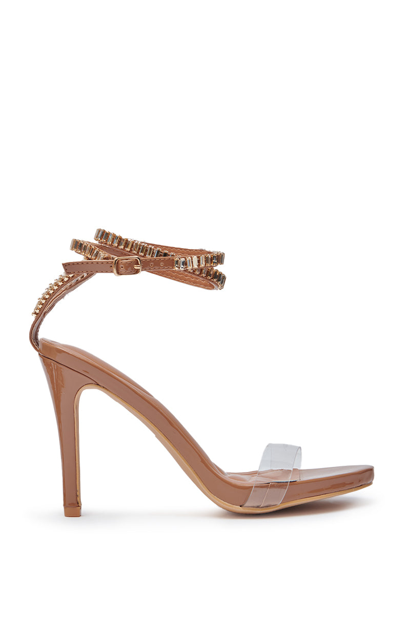 The AZALEA WANG ELLYIE TAN RHINESTONE STRAP SANDAL showcases a tan sole, stiletto heel, clear faux leather toe strap, and two gold-studded ankle straps with rhinestones. Displayed on a clean white background.