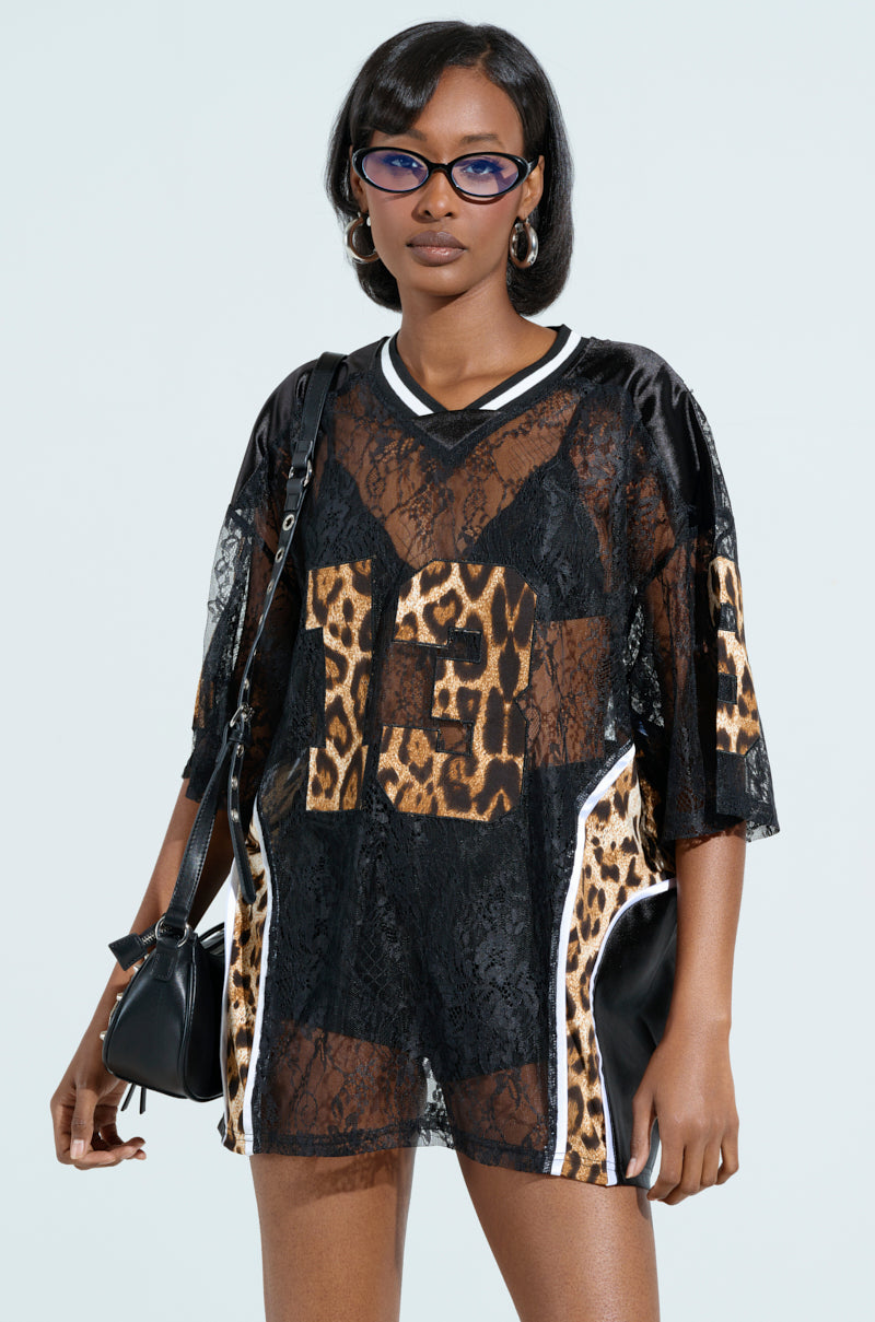 A woman confidently stands in a TAILGATE READY JERSEY, complemented by a black lace top with leopard print satin details. She accessorizes with a black handbag, stylish glasses, and hoop earrings against a plain light background.