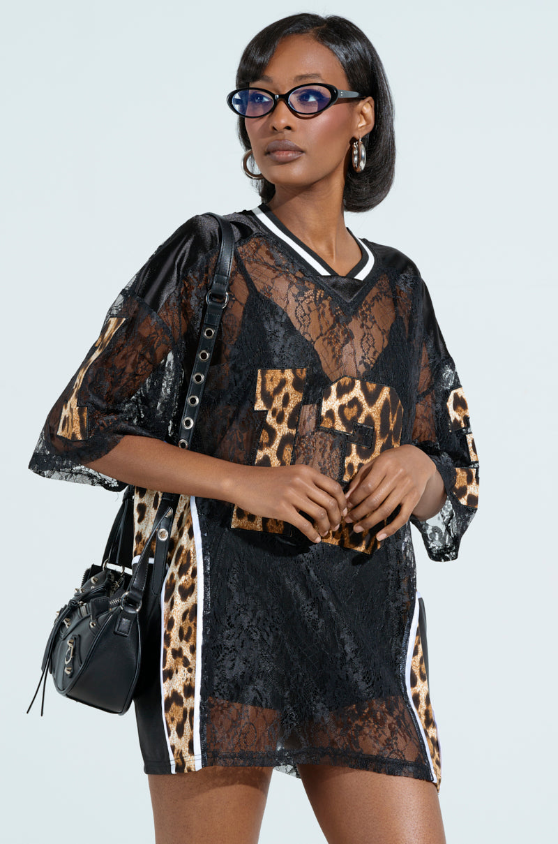 A person sporting blue glasses, hoop earrings, and short hair poses confidently in a black lace top over a TAILGATE READY JERSEY. They carry a black handbag against a light green background.