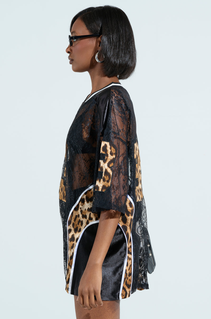 A person with short black hair, sunglasses, and hoop earrings exudes confidence in a stylish black lace dress with leopard print panels. Their look is polished off with a chic black handbag, perfectly matching the flair of the TAILGATE READY JERSEY against a light background.