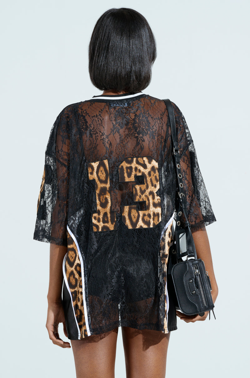 A person with shoulder-length black hair wears the TAILGATE READY JERSEY, a sheer black lace top with leopard print detailing and the number 13 on the back. They carry a black bag with a strap, standing against a light blue background.