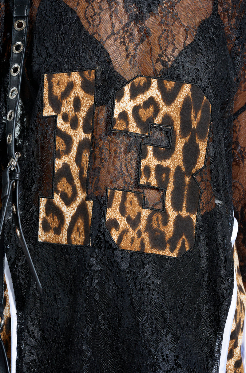 Close-up of a black lace fabric featuring a leopard print "13" design, reminiscent of a football top. A black strap with metal eyelets adorns the left side, offering a TAILGATE READY JERSEY feel by blending lace with dynamic animal print.