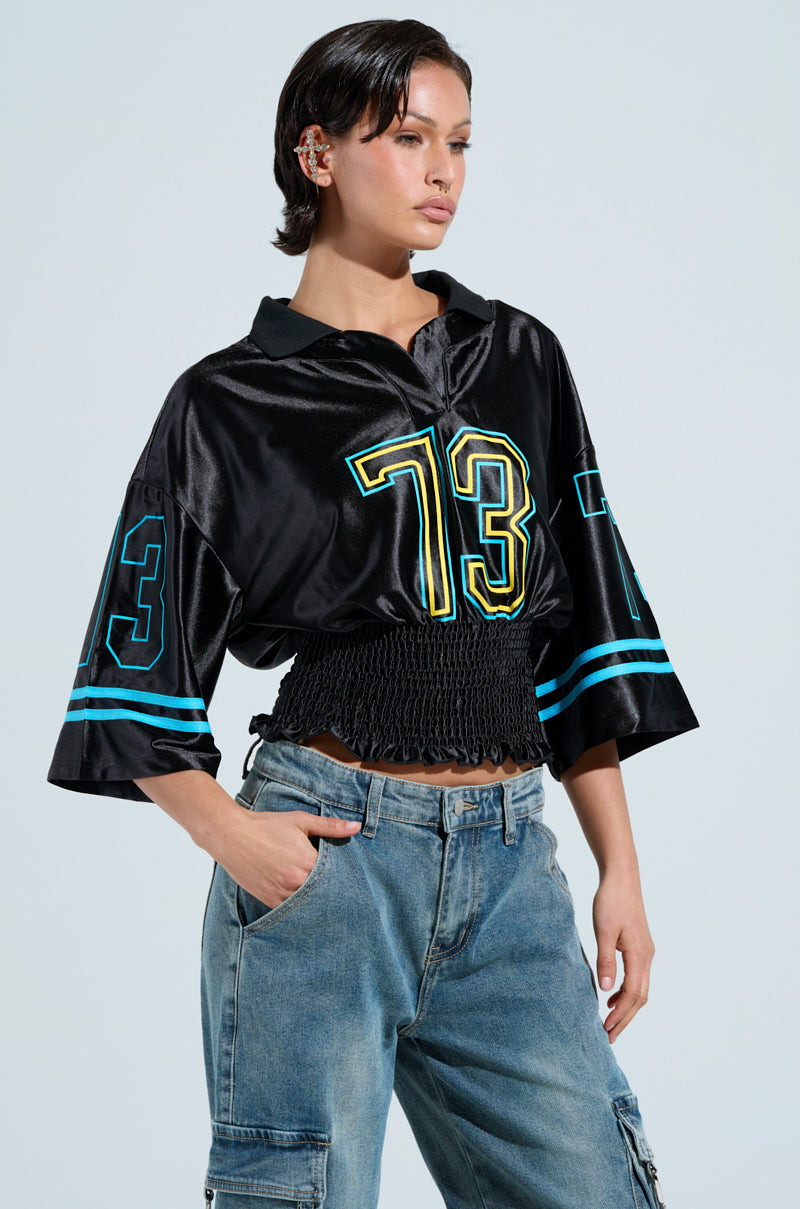 Someone confidently poses against a plain background wearing the CHAMPIONSHIP SCRUNCH JERSEY, featuring varsity-inspired stripes, the number 73, silky jersey fabric, and wide sleeves, paired with blue jeans.