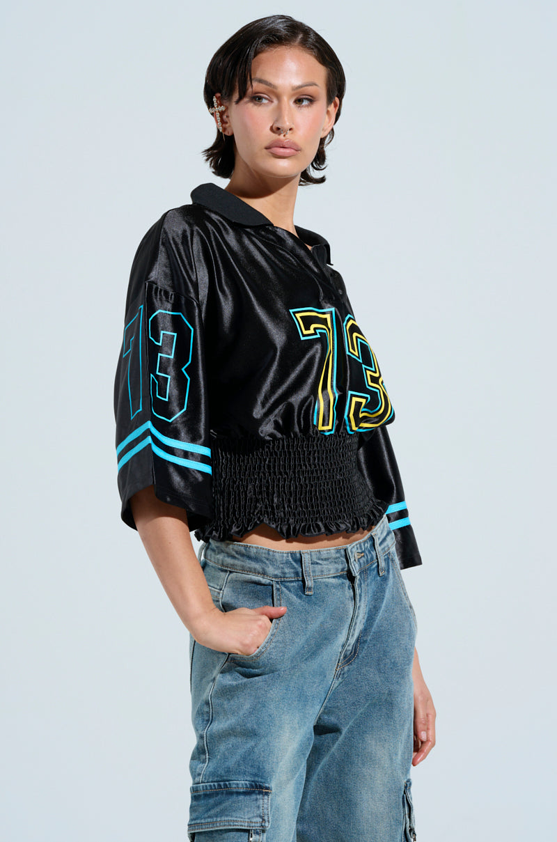 Wearing a confident smile, someone sports the silky CHAMPIONSHIP SCRUNCH JERSEY with number 73 in yellow and blue, paired with relaxed blue jeans. The varsity-inspired stripes stand out against the plain background, highlighting their casual yet stylish look.
