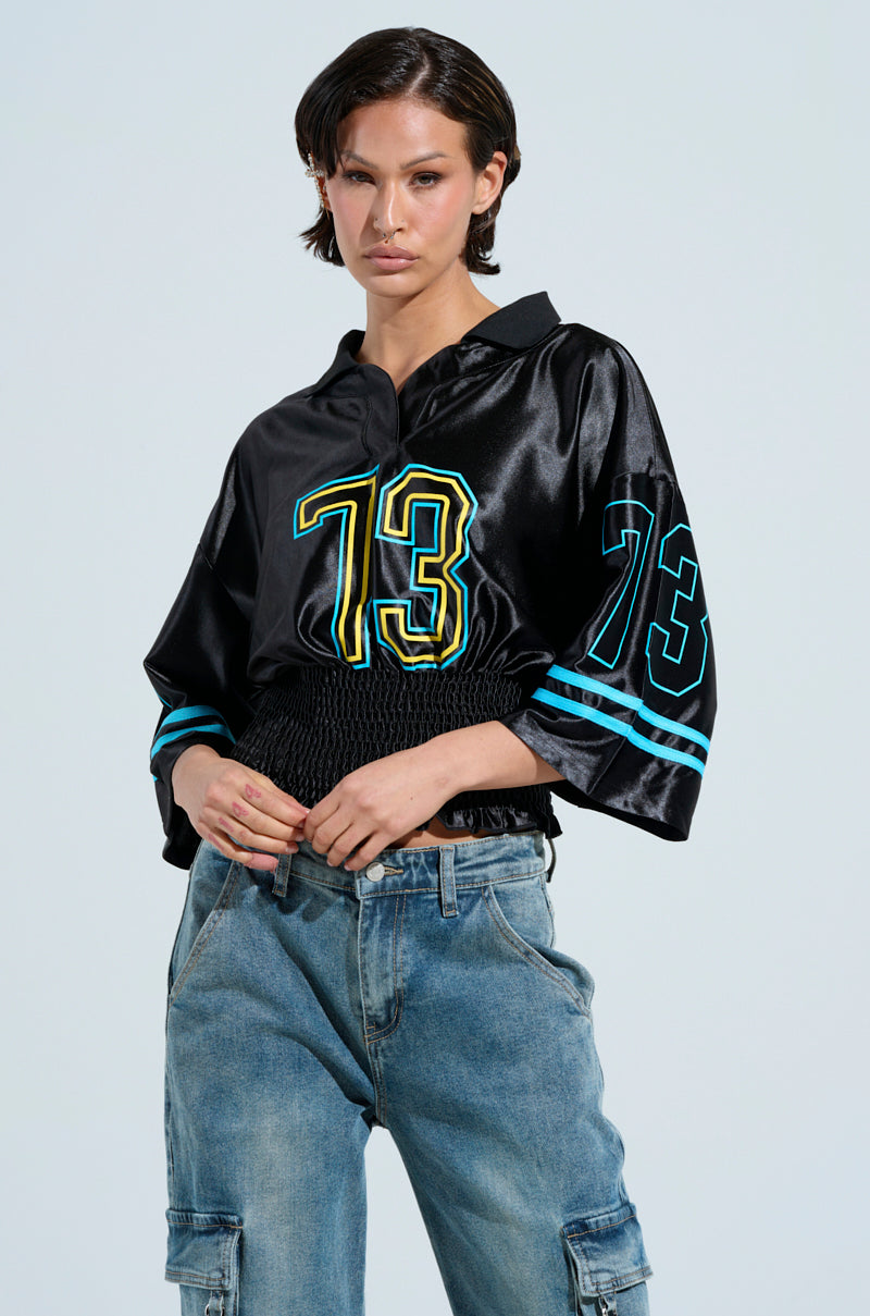 A person with short dark hair confidently wears a shiny black Championship Scrunch Jersey, featuring the number 73 in turquoise and yellow on both the front and sleeves. They pair it with casual light blue jeans against a light background.