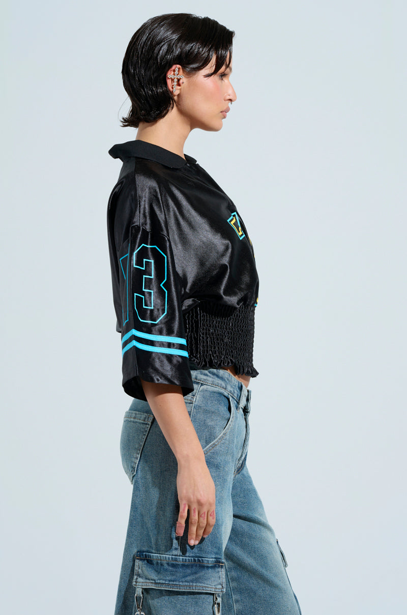 With short hair, the individual wears a black CHAMPIONSHIP SCRUNCH JERSEY with varsity stripes and the number 23 in silky fabric, plus blue accents. Paired with blue cargo jeans, they pose in profile against a plain grey backdrop.