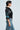 With short hair, the individual wears a black CHAMPIONSHIP SCRUNCH JERSEY with varsity stripes and the number 23 in silky fabric, plus blue accents. Paired with blue cargo jeans, they pose in profile against a plain grey backdrop.