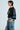 With short hair, a person in profile wears the CHAMPIONSHIP SCRUNCH JERSEY in black, featuring varsity-inspired stripes and the number 73. They pair it with denim cargo pants, hands in pockets. The silky jersey fabric stands out against the plain light blue background.