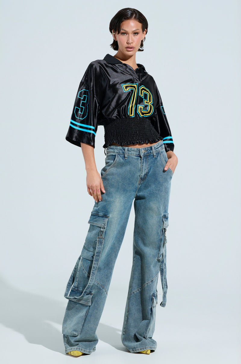 A person in a CHAMPIONSHIP SCRUNCH JERSEY with the number 73 and blue accents pairs it with baggy denim cargo pants. Varsity-inspired stripes enhance their style as they pose confidently against a plain background.