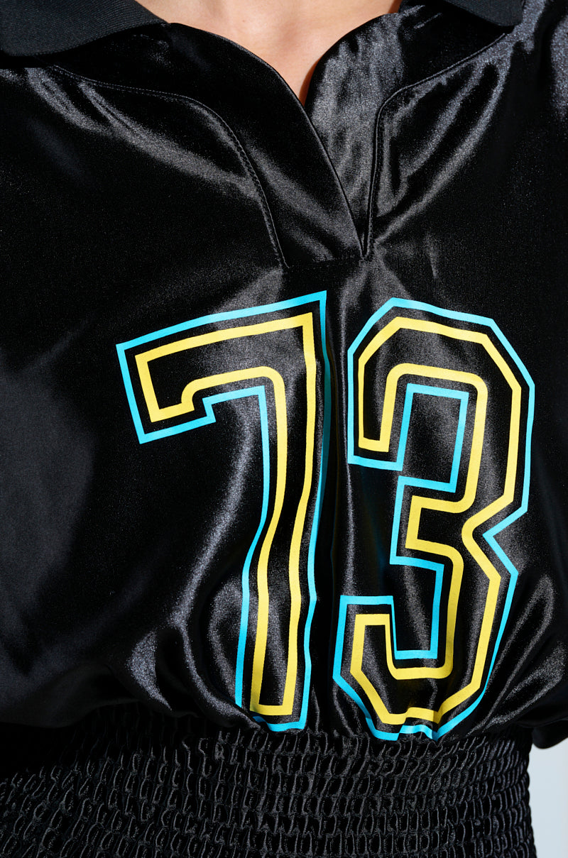 A person models the CHAMPIONSHIP SCRUNCH JERSEY, showcasing its shiny black design with bold number 73 outlined in yellow and blue. The jersey includes varsity-striped high collar and textured waist fabric.