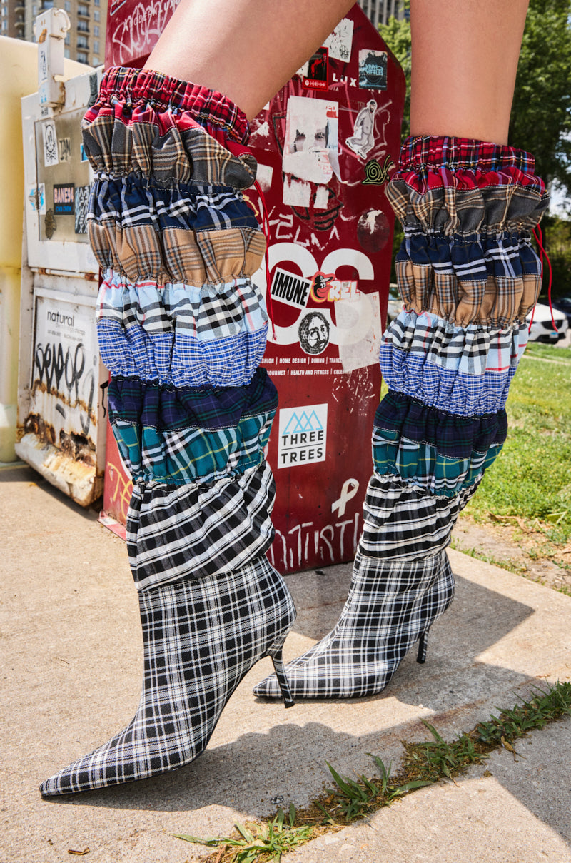 Wearing the AZALEA WANG ELODIE MULTI BOXER BOOT—knee-high, plaid, and made from layers of colorful fabric—a person stands by a red sticker-covered utility box and green grass.