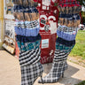 Wearing the AZALEA WANG ELODIE MULTI BOXER BOOT—knee-high, plaid, and made from layers of colorful fabric—a person stands by a red sticker-covered utility box and green grass.